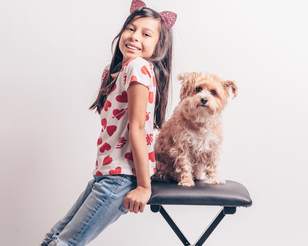 A girl leans on the stool her dog is perched on