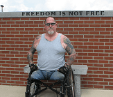 A veteran who lost his legs sitting in a wheelchair in front of a stone wall that reads, freedom is not free